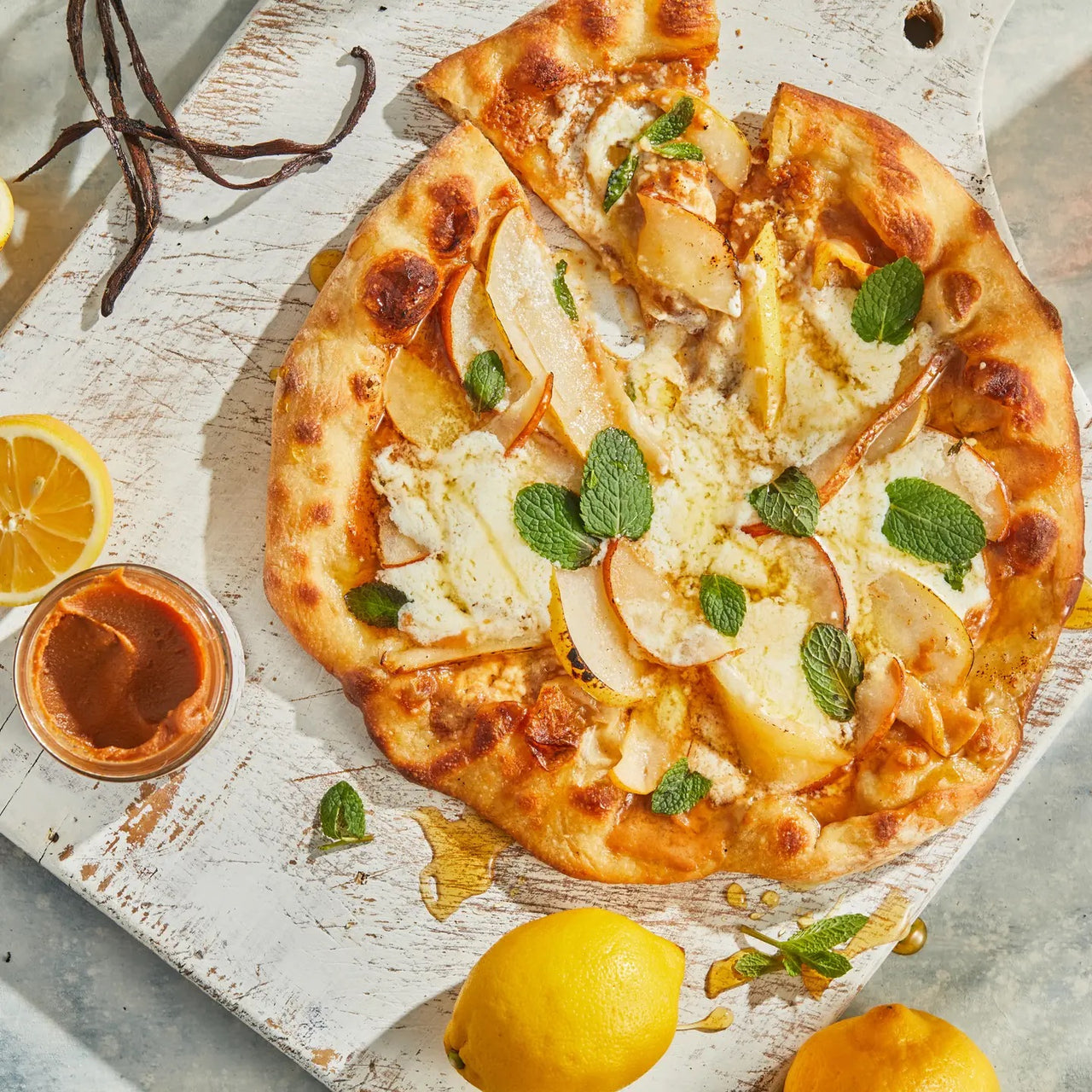 A rustic pizza with ricotta, pear, fresh mint, and Lemon Vanilla Honey Cream Spread 🍋 – Tart, Sweet & Totally Irresistible sits on a white wooden board with lemons, vanilla beans, and a dish of spice paste.