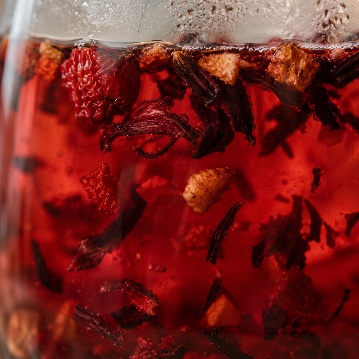 Close-up of a glass filled with a deep red beverage or tea, suspended with dried botanical pieces and fruit pieces visible throughout.