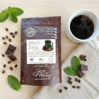 Thumbnail for Minty Thins coffee bag with mug of black coffee, mint leaves, chocolate pieces, and coffee beans
