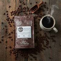 Thumbnail for A bag of Java Momma Indian Monsoon Malabar Coffee | Dark Roast, Low Acid, Single Origin rests on a wooden surface, surrounded by scattered beans, a steaming mug of black coffee, and a small wooden scoop filled with beans.