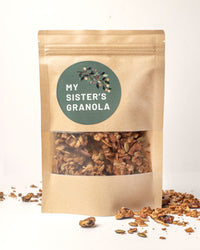 Thumbnail for A brown resealable pouch with a clear window displays My Sister’s Granola – Nutty, Sweet, and Totally Addictive on a green label with a branch design. Small batch granola pieces are scattered around the bag.