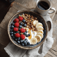 Thumbnail for A rustic ceramic bowl with yogurt, blueberries, raspberries, banana slices, My Sister’s Granola – Nutty, Sweet, and Totally Addictive, toasted coconut, and honey sits beside a cup of black coffee on a wooden table.