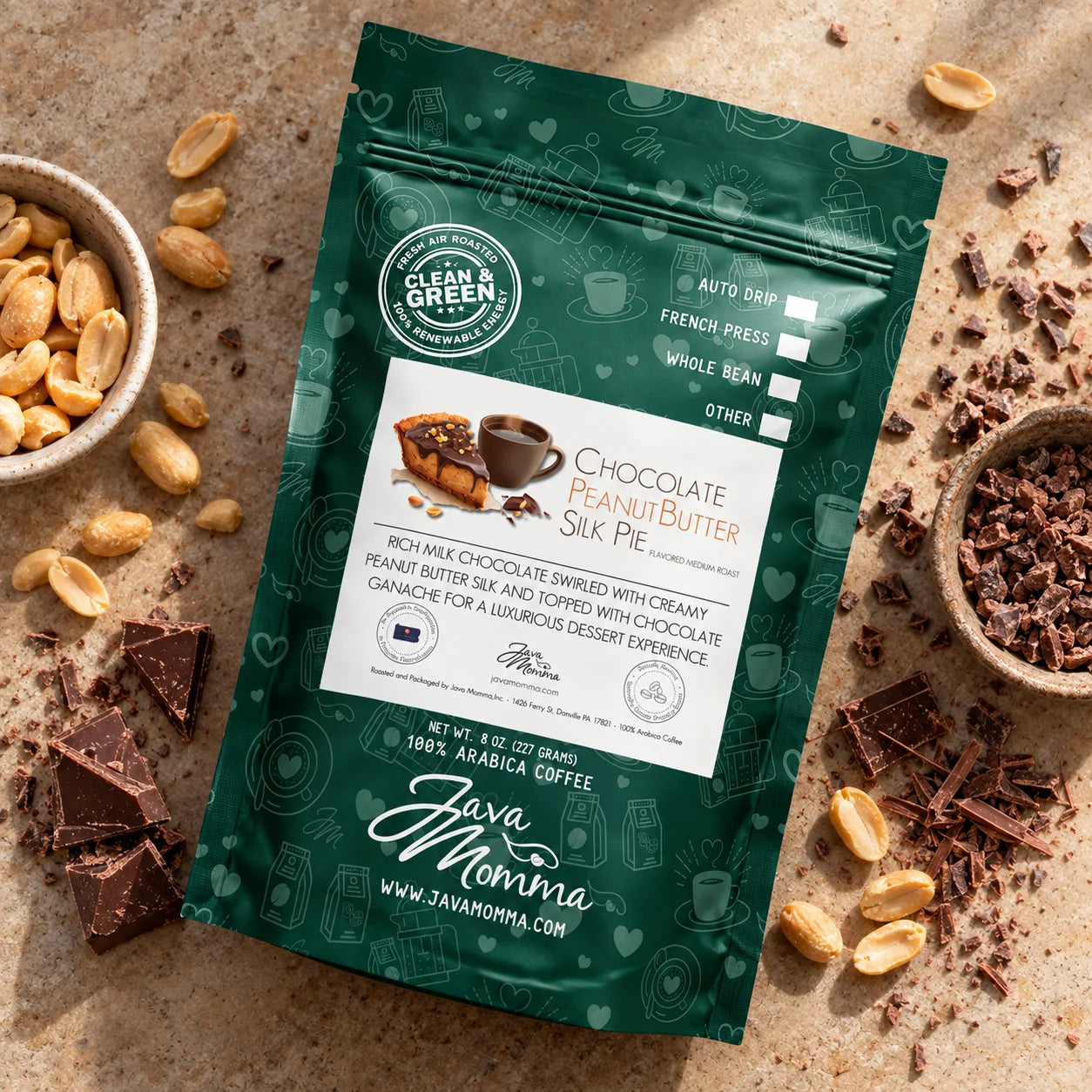 A stand-up pouch of Java Momma Chocolate Peanut Butter Silk Pie Arabica coffee is displayed on a textured beige stone surface, surrounded by chocolate pieces, whole peanuts, and chocolate crumbs. The packaging is dark green with a white central label and features the Java Momma brand logo.