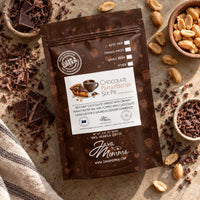 Thumbnail for A stand-up coffee pouch labeled 'Chocolate Peanut Butter Silk Pie' by Java Momma is photographed from above on a textured stone surface, surrounded by chocolate pieces, peanuts, and dried coffee beans. The package is dark brown with a large white front label and small coffee-themed illustrations.