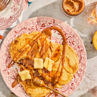 Thumbnail for A plate of pancakes topped with apples, caramel sauce, honey, and butter sits on a pink floral plate. Beside it is a jar of Peach Vanilla Honey Cream Spread 🍑 – Sweet Summer Bliss in a Jar and a glass of refreshing drink.