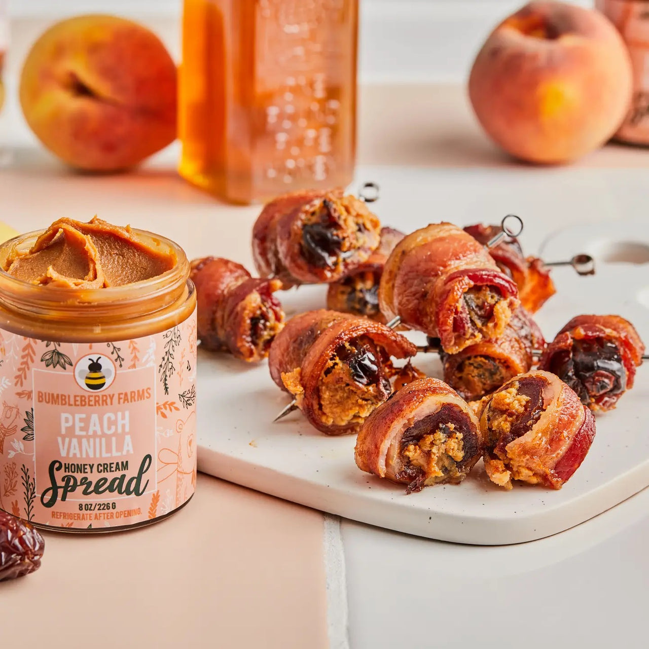 A jar of Peach Vanilla Honey Cream Spread 🍑 – Sweet Summer Bliss in a Jar sits beside sliced peach and bacon-wrapped stuffed dates on a white cutting board, showcasing this creamy blend of peaches, vanilla, and honey.
