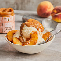 Thumbnail for A bowl of vanilla ice cream with caramel sauce and sliced peaches, served with a spoon. In the background sits Peach Vanilla Honey Cream Spread 🍑 – Sweet Summer Bliss in a Jar, alongside whole peaches and a metal scoop.