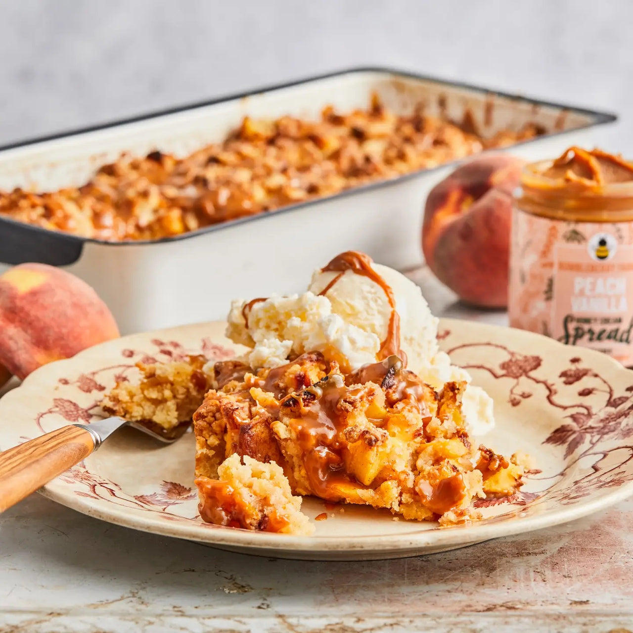 A plate of peach cobbler topped with vanilla ice cream and caramel sits beside a fork. In the background: a baking dish, peaches, and a jar of Peach Vanilla Honey Cream Spread 🍑 – Sweet Summer Bliss in a Jar.