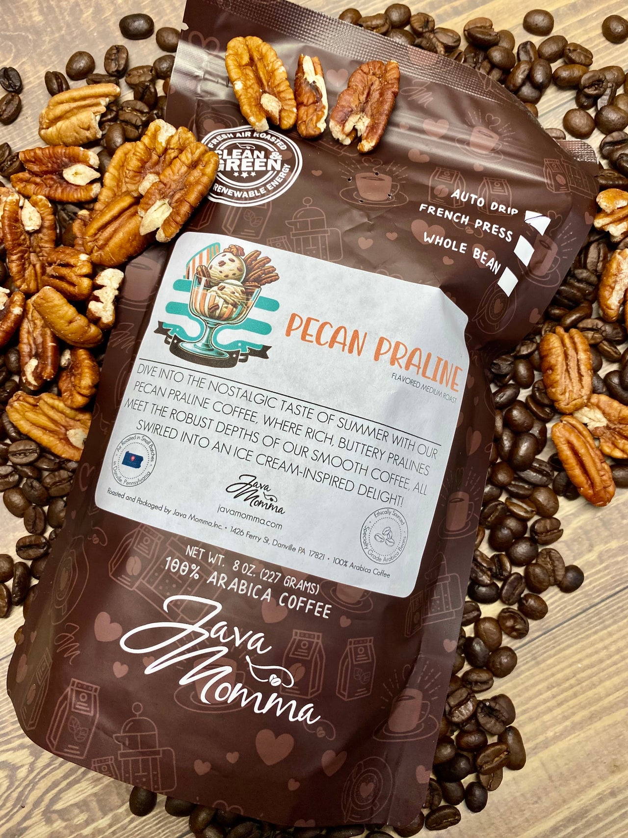 A bag of Java Momma Pecan Praline – a buttery, nutty medium roast inspired by a Southern summer treat – sits on wood, surrounded by beans and pecan halves. The brown package highlights smooth praline flavors and brewing tips.