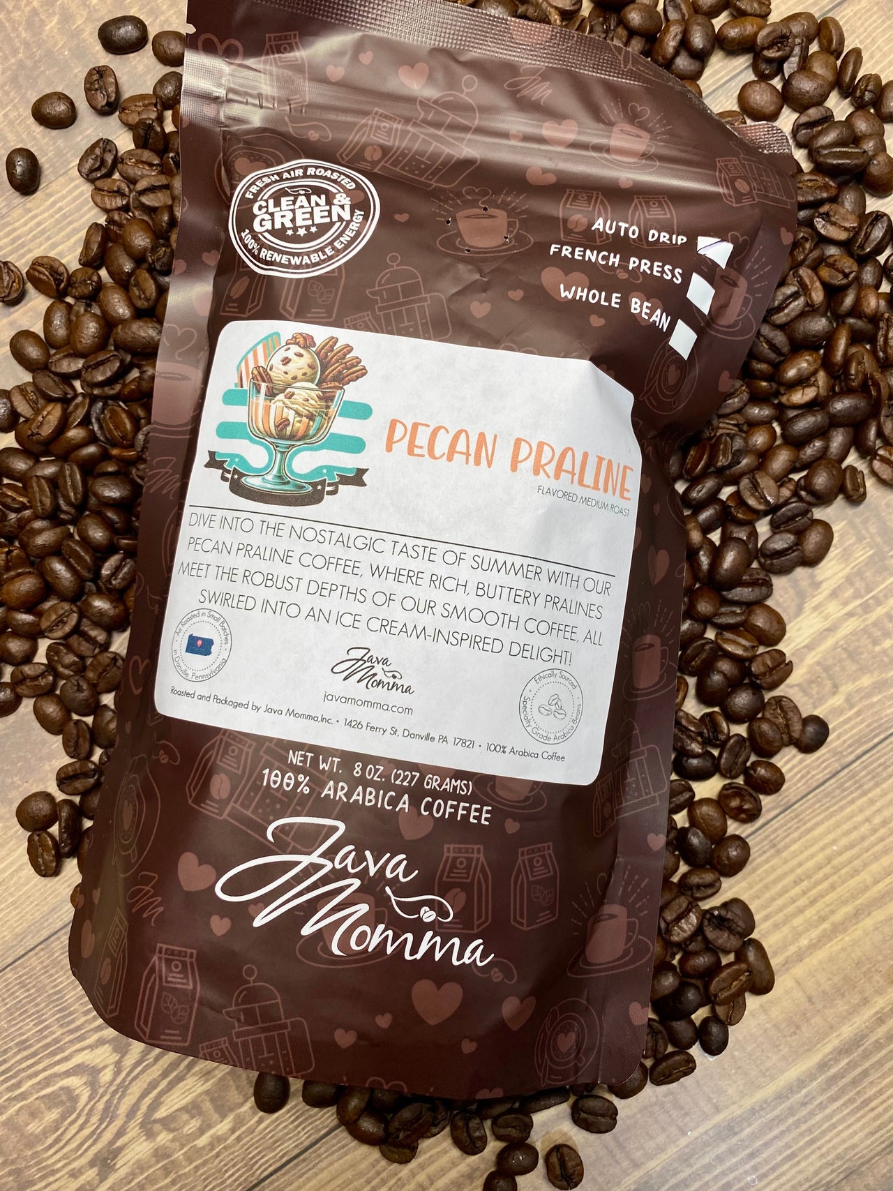 A bag of Java Momma Pecan Praline – a buttery, nutty medium roast inspired by a Southern summer treat and keto friendly – rests on a surface with scattered beans. The bag shows a coffee cup with pecans and an ice cream swirl.