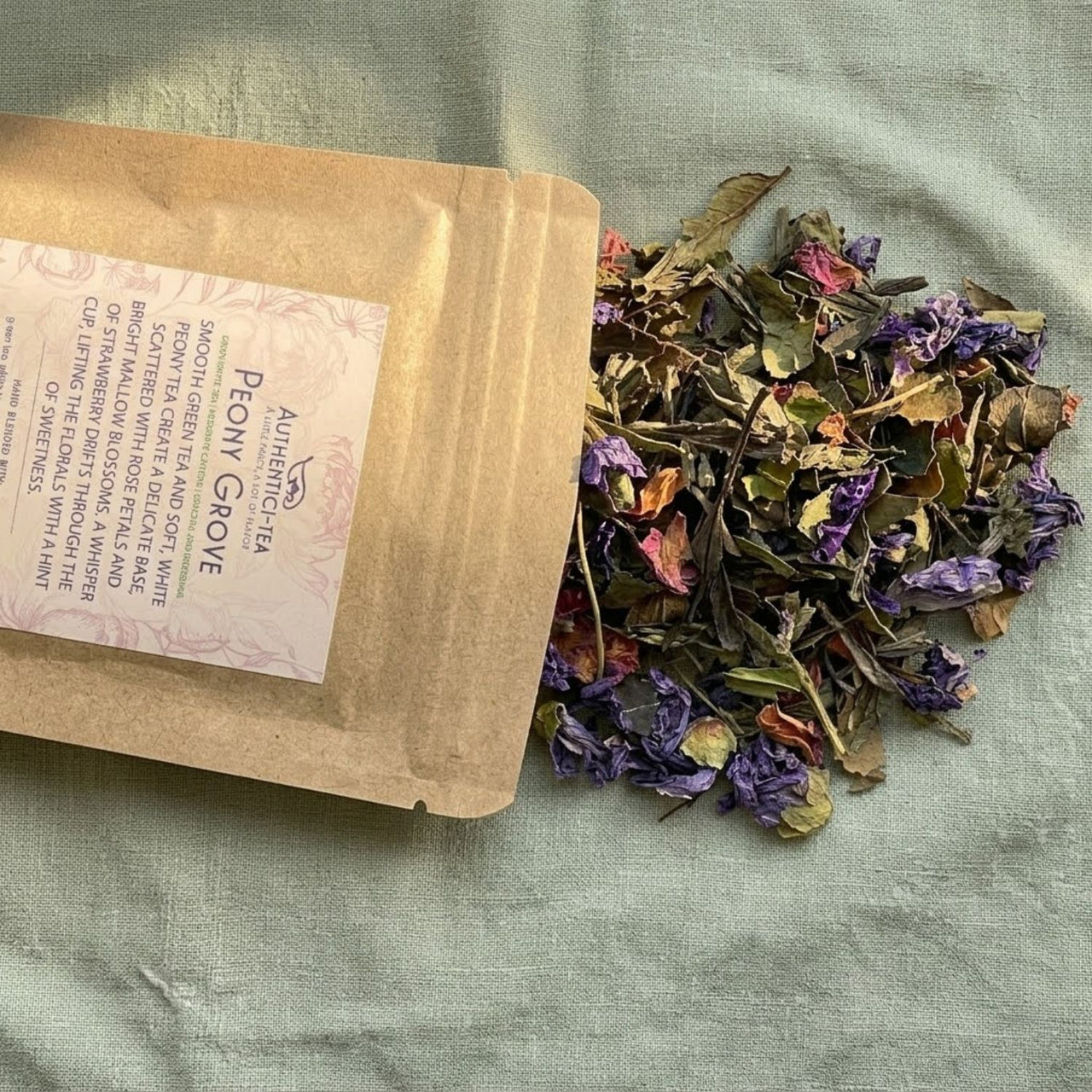 Peony Grove green & white tea blend package spilling tea leaves and dried flowers on a light fabric surface.