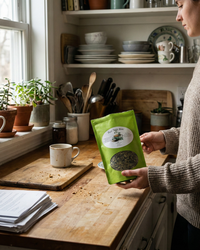 Thumbnail for Woman holding Plot Twist Mint Tea | Darjeeling & Peppermint Black Tea Blend in a cozy kitchen with a cup on the table.