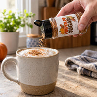 Thumbnail for A hand holds a clear spice/sugar shaker labeled 'Pumpkin Pie Pie Sugar' above a ceramic mug filled with a latte, sprinkling the seasoning over the foam. The mug is set on a kitchen counter with a plant, pumpkin, and towel in the background.