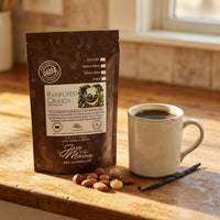 Thumbnail for Rainforest Crunch bag on a worn wood countertop beside a ceramic mug filled 
with coffee. Scatter a small handful of raw almonds, hazelnuts, and macadamia nuts nearby. 
One vanilla bean pod resting at the base of the mug. Warm morning-to-afternoon light. 
Cozy, lived-in kitchen feel.
