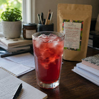 Thumbnail for A glass of iced red beverage with ice cubes sits on a wooden desk, with a kraft pouch labeled Raspberry Rebel behind it and notebooks, pens, and a plant in the background.