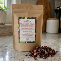 Thumbnail for A stand-up kraft paper pouch of Authentic Tea Company's Raspberry Rebel loose-leaf tea sits on a speckled granite countertop with a small pile of the loose tea in front. The background shows a softly blurred kitchen setting.