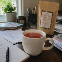 Thumbnail for A steaming ceramic mug filled with raspberry iced tea sits on a wooden desk next to a pouch of Raspberry iced Tea. The scene includes a notebook, pen, and other desk items in a cozy indoor setting.