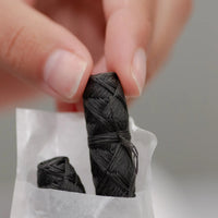Thumbnail for A hand holds a small black bundle of No Tox Life® Charcoal Floss Refills – 2-Pack, with another refill on white paper below—an eco-friendly, gentle-on-gums choice for low-waste dental care.