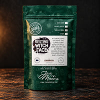 Thumbnail for A green stand-up pouch of Java Momma 'Resting Witch Face+' coffee on a dark wooden surface against a dark background. The packaging features white text, coffee-themed illustrations, and a circular 'Clean Green Certified' seal.