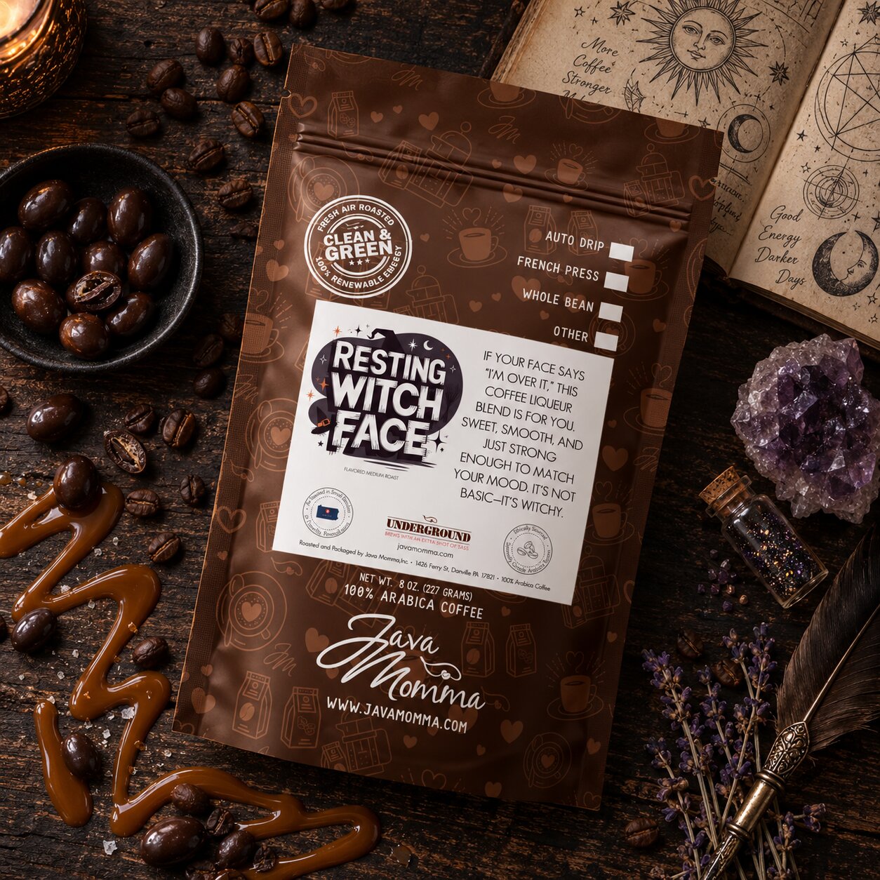 A bag of Java Momma Coffee by Java Momma is displayed on a dark wooden table surrounded by coffee beans, caramel drizzle, an amethyst crystal, and an open sketchbook.