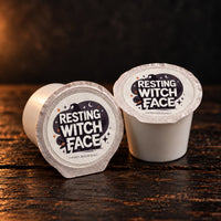 Thumbnail for Two coffee pods from Resting Witch Face on a dark wooden surface with a dark background. The pods are white with silver foil lids and a black label featuring a witch hat and pumpkin graphic.