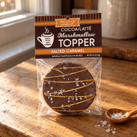 Thumbnail for A packaged round chocolate topper on a wooden tabletop near a window. The product is labeled Melville Cocoa/Latté Marshmallow Topper, Salted Caramel, and is shown in a clear bag with a brown header card.