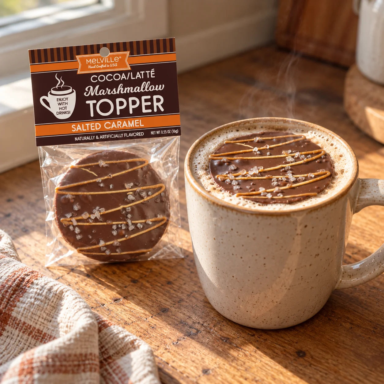 A packaged chocolate marshmallow topped with caramel and coarse salt sits beside a ceramic mug filled with a hot cocoa drink on a wooden table.
