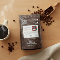 Thumbnail for A bag of Smooth Operator Mixed Roast Blend | Low Acid Coffee sits on a wooden table with coffee beans, a steaming mug, a scoop, and a beige cloth—ideal for those seeking gentle coffee for sensitive stomachs.