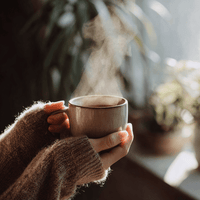 Thumbnail for A person in a cozy sweater holds a steaming mug of Smooth Operator Mixed Roast Blend | Low Acid Coffee. Soft sunlight filters through blurred plants behind them, creating a warm and comforting atmosphere.