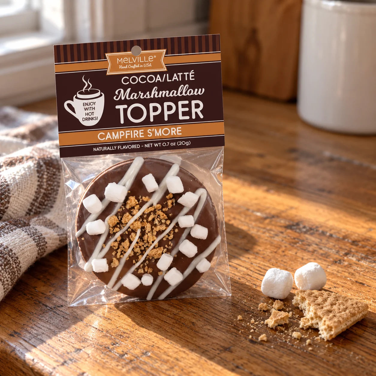 Melville Cocoa Latté Marshmallow Topper Campfire S'more in a clear bag with the branded header card, photographed on a wooden table with a graham cracker and marshmallow nearby.