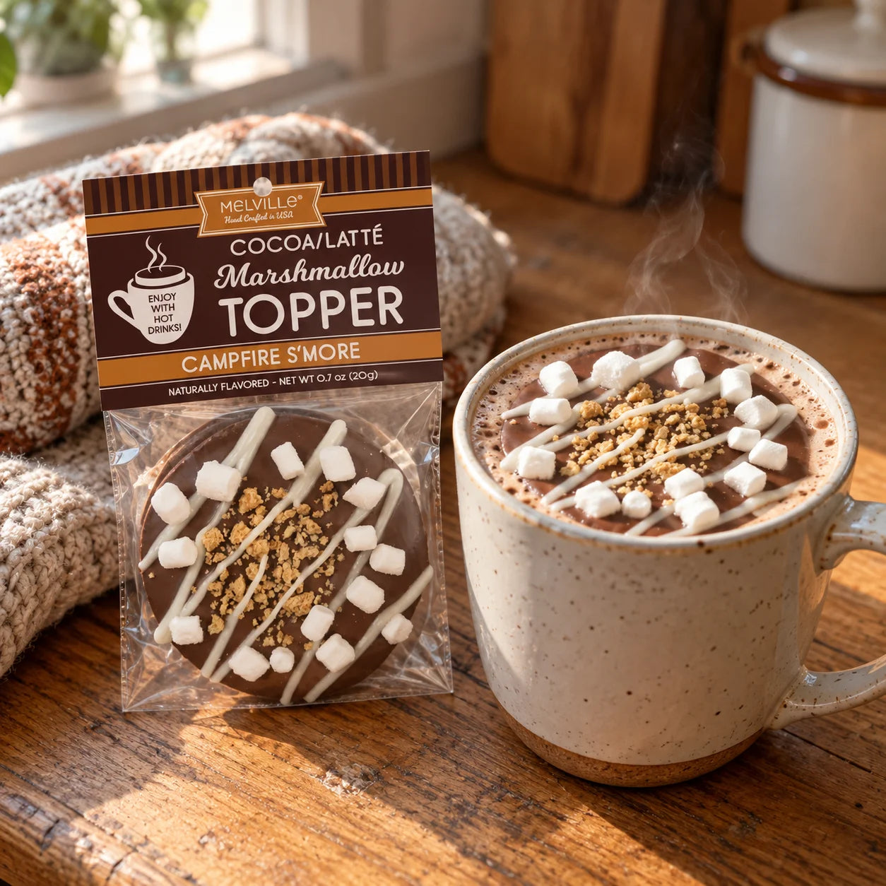 A packaged Melville Cocoa/Latté Marshmallow Topper in a campfire s'more flavor, shown next to a ceramic mug filled with hot cocoa topped with marshmallows, nuts, and drizzle on a wooden table.
