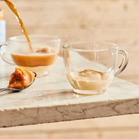 Thumbnail for A spoonful of Sea Salt 🧂 Caramel Honey Cream Spread – Sweet, Salty, & Sinfully Smooth rests on marble next to a glass of cream, with coffee being poured into another glass cup in the background.