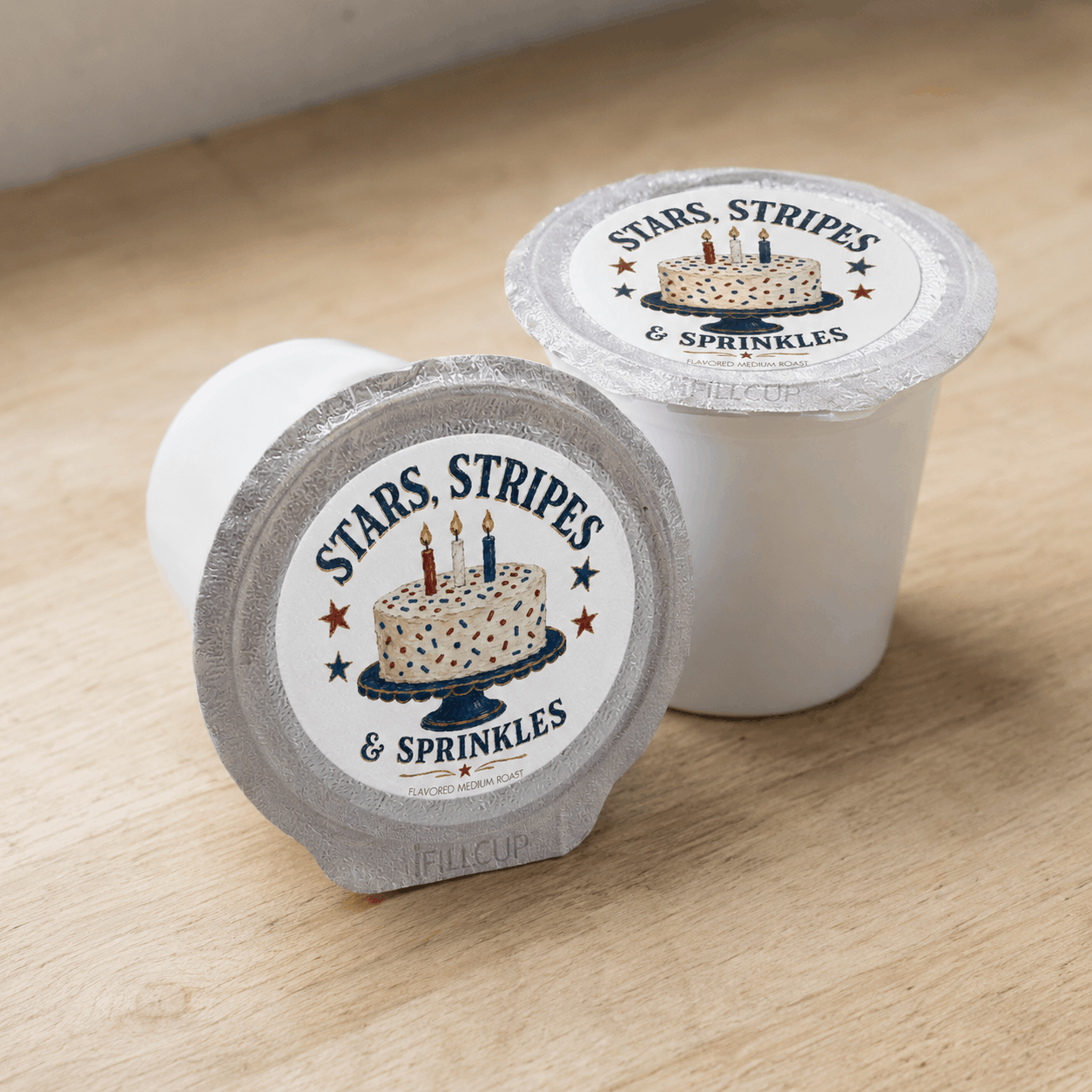 Two Stars, Stripes & Sprinkles single-serve coffee pods on a wood surface, showing the folk art birthday cake label with navy, red, and cream Americana design.