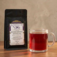 Thumbnail for Black Authentic-Tea Strawberry Serendipity pouch beside a clear glass mug of red herbal tea on a wooden table with soft steam rising.