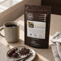Thumbnail for Java Momma Sumatra Extra Dark coffee bag next to a mug of coffee with cherries, cacao nibs, and a vanilla bean pod
