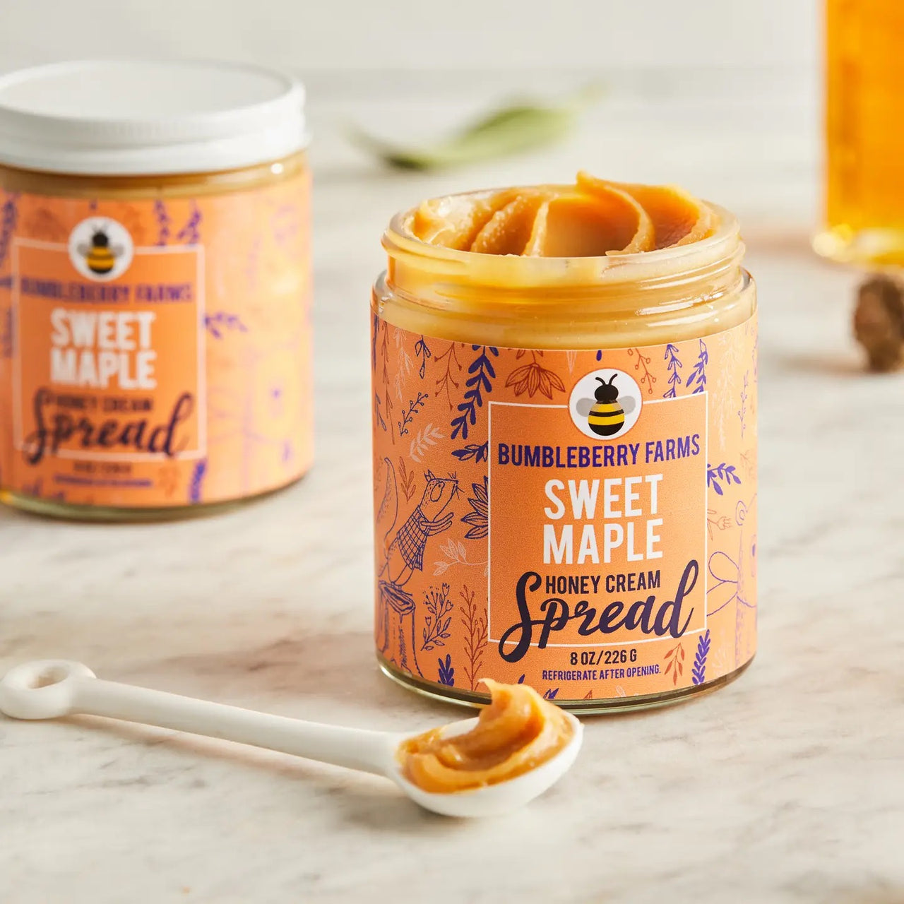 An open jar of Sweet Maple 🍁 Honey Cream Spread – Cozy Maple Bliss in Every Bite sits on a marble surface with a spoonful beside it, showcasing the rich blend of maple syrup and wildflower honey; another closed jar is behind.