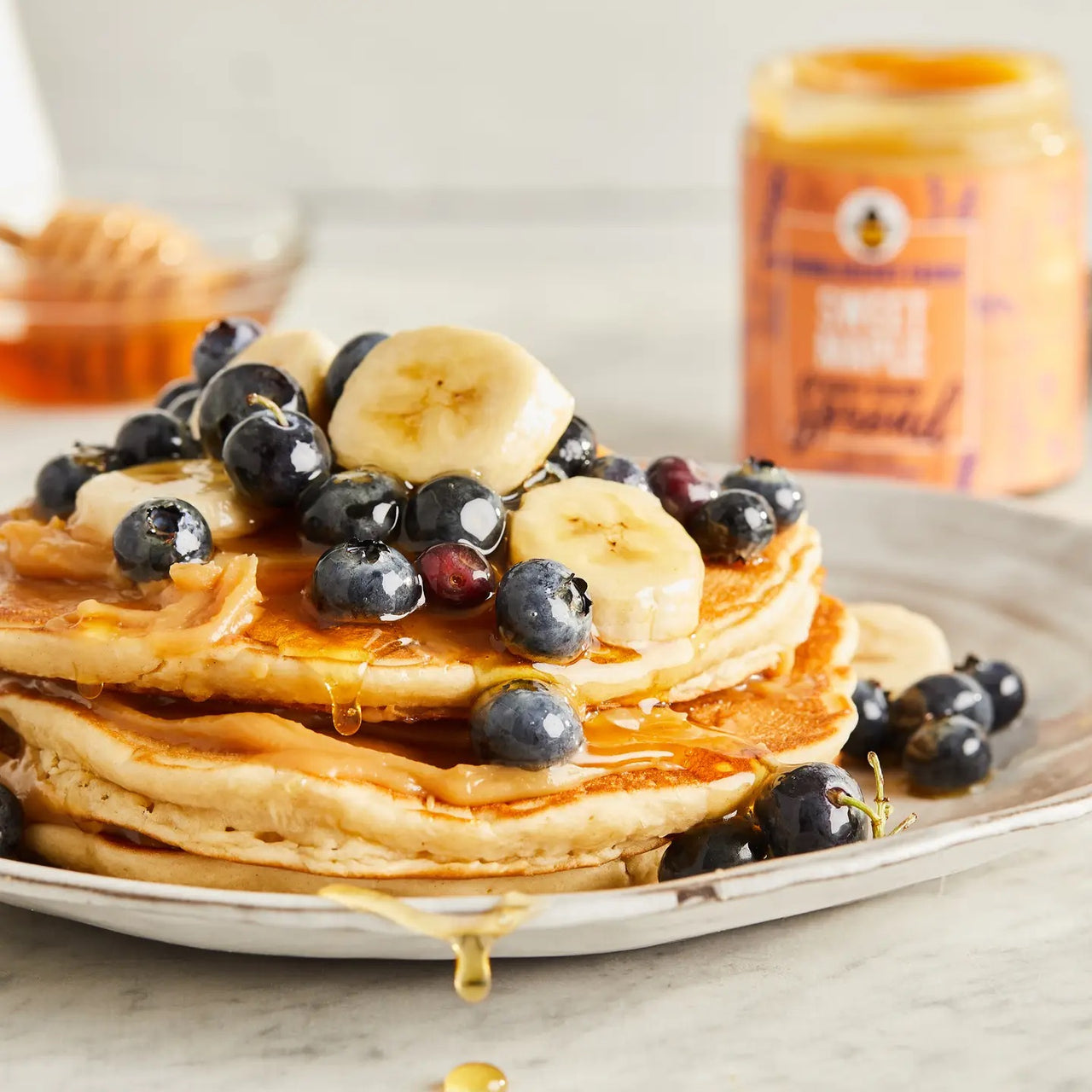 A stack of pancakes topped with banana slices, blueberries, and a drizzle of Sweet Maple 🍁 Honey Cream Spread sits on a plate. In the background, a honey dipper and a jar of spread are softly blurred.