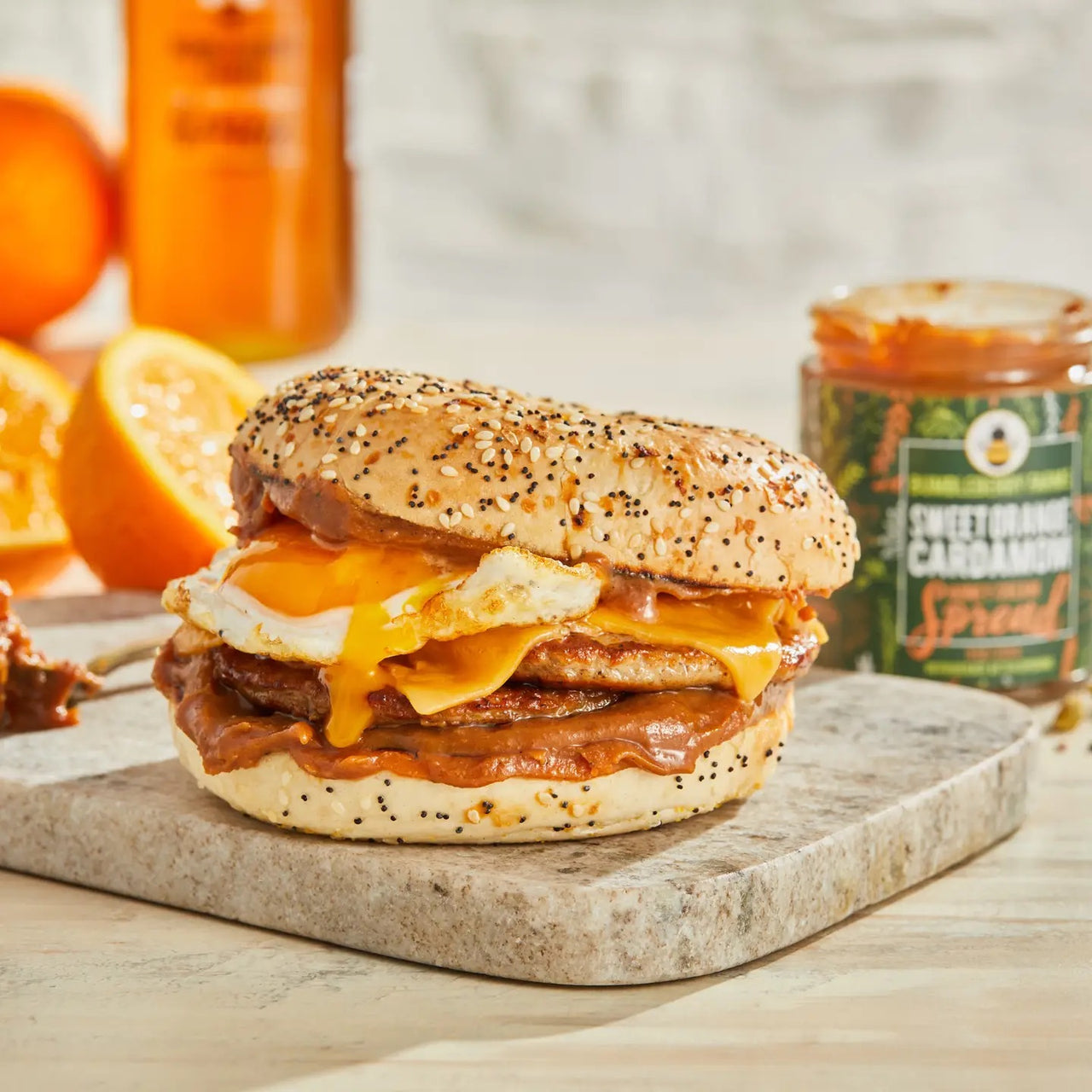 A seeded bagel sandwich with fried egg, melted cheese, and Sweet Orange Cardamom Honey Cream Spread 🍊 sits on a stone board. Behind it are orange slices, a jar of the spread labeled “Bold Citrus Meets Cozy Spice,” and a bottle.