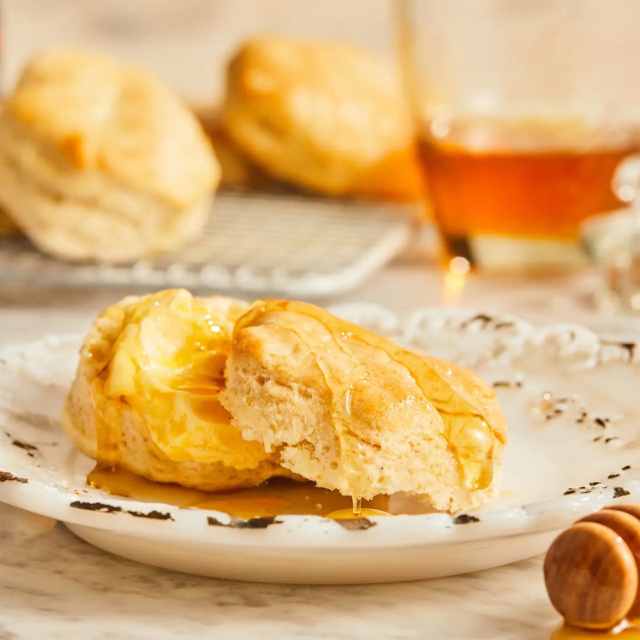 A flaky biscuit split open with butter and Sweet Orange Cardamom Honey Cream Spread 🍊 sits on a white plate. More biscuits and a jar of the citrus-spice honey spread are blurred in the background.