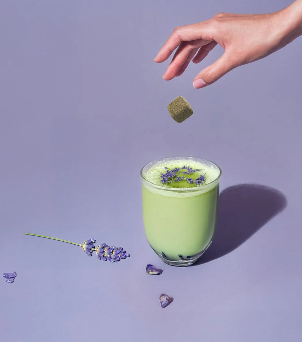 A hand drops a Lavender Matcha Tea Drop – Calm Energy in Every Sip 💜🍵 into a glass, garnished with lavender flowers on a purple surface, with a sprig of lavender nearby, showcasing vibrant organic matcha.