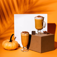 Thumbnail for Two glasses of pumpkin spice latte, made with the Pumpkin Spice Latte Kit – Fall’s Coziest Tea Latte Gift Set 🎃☕🍁 by Tea Drops, are topped with whipped cream and cinnamon beside a small pumpkin, chocolate cube, and cinnamon sticks on an orange background.