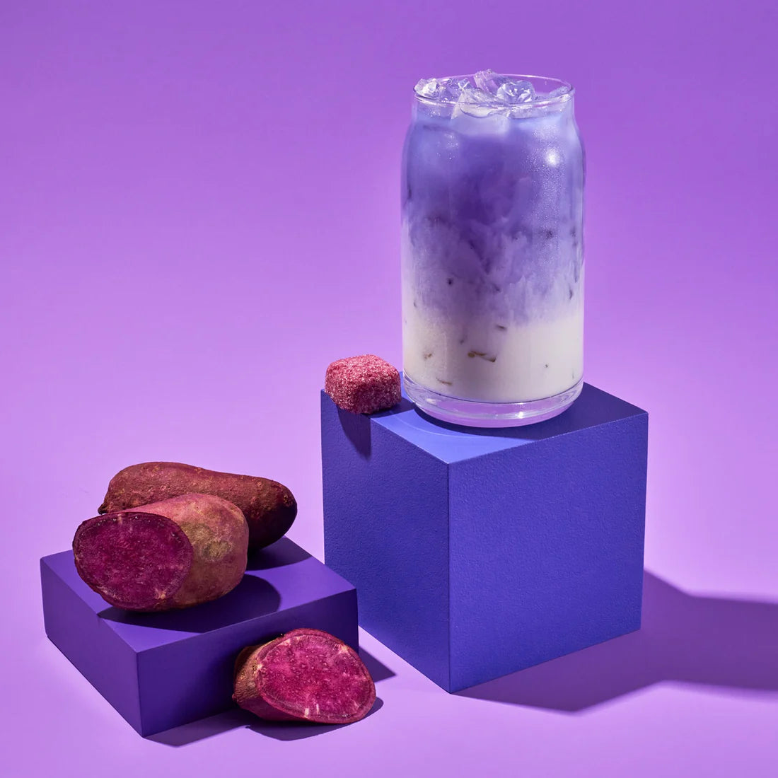 A glass of iced, purple and white layered 💜 Ube Tea Drops® – A Sweet, Nutty, & Creamy Purple Delight! 🍠✨ sits on a purple block with whole and sliced purple sweet potatoes, all set against a matching purple background. Caffeine free!.