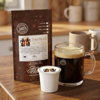 Thumbnail for Three Beans Butter Beer coffee bag with single serve pod and glass mug of freshly brewed coffee with light foam on a warm wood kitchen counter.