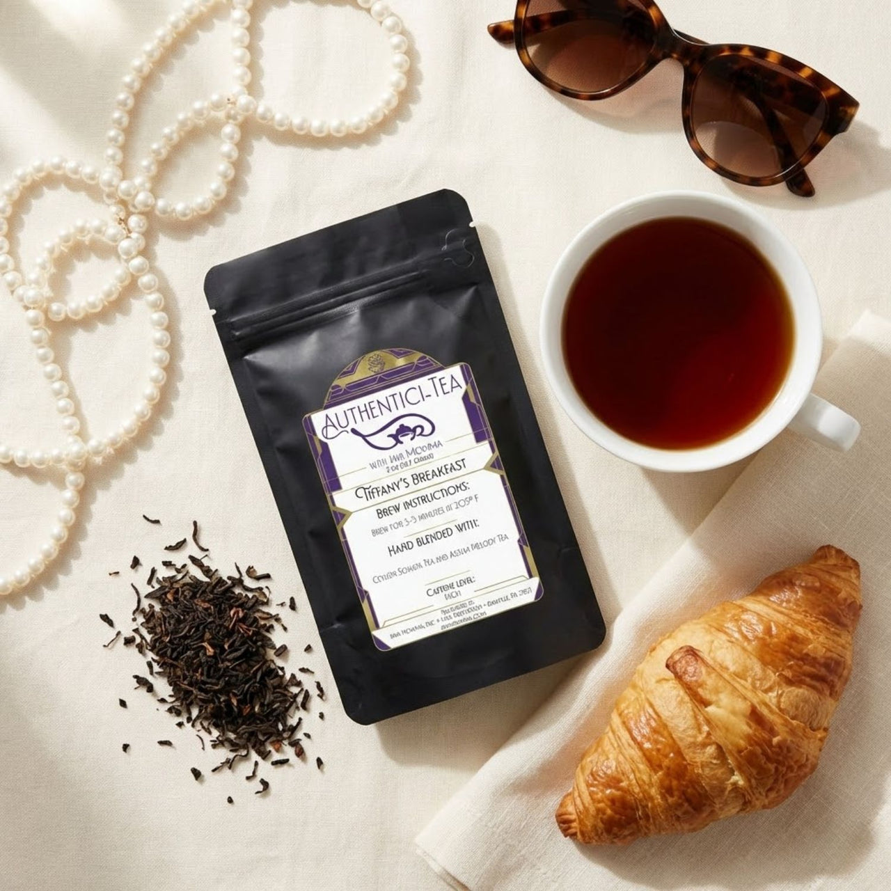 Tiffany's Breakfast Tea packaging with a cup of tea, croissant, and loose tea leaves on a light surface.